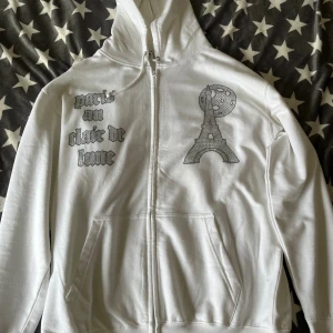 Blinglucid vit hoodie - Vit hoodie med dragkedja från Blinglucid, dekorerad med glittrande stenar som bildar texten 'paris au clair de lune' och motiv av Eiffeltornet och månen. Tröjan har huva med snörning och två fickor framtill. Perfekt för dig som gillar statement-plagg och coola detaljer.