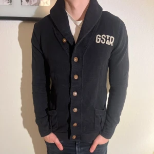 Svart kofta från G-Star RAW - Svart kofta från G-Star RAW med broderad logga på bröstet. Koftan har stora bruna knappar, två fickor framtill och en bred krage. Tillverkad i ett stickat bomullsmaterial som känns mjukt och bekvämt. Perfekt för lager-på-lager under kyliga dagar. Sönder vid lilla taggen på sidan av tröjan