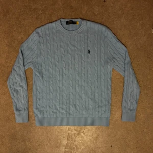 Ljusblå Polo Ralph Lauren - Snygg ljusblå stickad tröja från Polo Ralph Lauren med klassiskt kabelstickat mönster och broderad logga på bröstet. Rund halsringning och ribbade muddar vid ärmslut och nederkant. Perfekt för dig som gillar stilrena och tidlösa plagg.