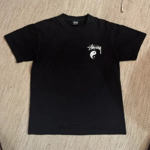  Stüssy t-shirt - Svart t-shirt från Stüssy i bomull med stor vit yin yang-tryck och logga på ryggen samt mindre tryck på bröstet. Klassisk rund hals och korta ärmar. Perfekt för dig som gillar streetwear och vill sticka ut med en fet grafisk design.
