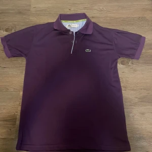 Lila pikétröja från Lacoste M - Snygg lila pikétröja från Lacoste med klassisk krage och knappar framtill. Tröjan har korta ärmar och den ikoniska krokodilloggan broderad på bröstet. Perfekt för en clean och sportig look.