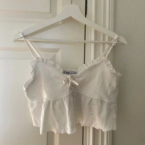 Vit linnetopp med spets från FB Sister - Superfin vit linnetopp från FB Sister med smala axelband, volangkant och broderade spetsdetaljer. Toppen har en söt knytning framtill och är croppad med lätt rynkad struktur. Perfekt för sommaren. 