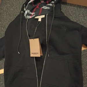 Svart Burberry hoodie med dragkedja - Svart hoodie från Burberry i storlek M med klassisk rutmönstrad huva i svart, vit och röd. Tröjan har snörning vid huvan, två fickor fram och snyggt Burberry-broderi på bröstet. Materialet är mjuk bomull och dragkedjan framtill ger en clean look.