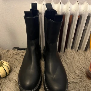 Svarta boots - Svart boots, platt sula och vita kontrastsömmar runt sulan. Skorna har dragflikar fram och bak för enkel påtagning och är tillverkade i ett matt skinnliknande material. Dem e använda 1 gång sen har dem bara stått så dem e som nya 