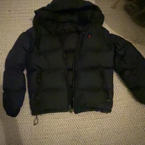 Svart dunjacka från Polo Ralph Lauren - Svart dunjacka från Polo Ralph Lauren med huva och dragkedja framtill. Jackan har puffiga sektioner, två sidofickor med dragkedja och en liten röd Polo-logga på bröstet. Perfekt för kalla dagar och har en clean, sportig look.