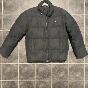 Svart pufferjacka från GANT - Svart pufferjacka från GANT med hög krage och dragkedja framtill. Jackan har två fickor med tryckknappar och en diskret logga på bröstet. Perfekt för kalla dagar och har en clean, stilren look som funkar till det mesta.                                           -Finns en luva som man kan sätta på 