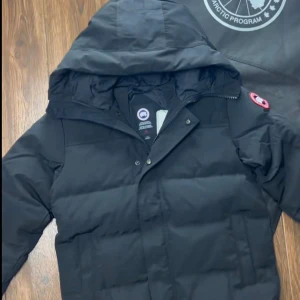 Svart dunjacka från Canada Goose - Säljer en svart dunjacka från Canada Goose med huva och klassisk logga på ärmen. Jackan har dragkedja och tryckknappar framtill samt praktiska axelremmar på insidan. Perfekt för kalla vinterdagar.