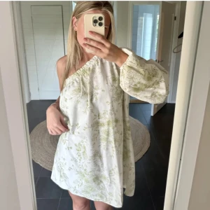 Vit blommig oneshoulder klänning - Säljer en vit kortklänning med ljusgröna blommor och oneshoulder-design. Klänningen har en lång puffärm och avslappnad passform, perfekt för sommarens varma dagar. Materialet känns lätt och luftigt, och den blommiga stilen ger en fräsch vibe.