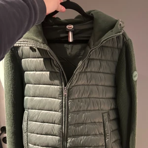 Grön cardigan Colmar Originals - Snygg grön pufferjacka från Colmar Originals med stickade ärmar och quiltad kropp. Jackan har huva, dragkedja framtill och två fickor. Märkeslogga på ärmen och cool blandning av material som ger en modern vibe.