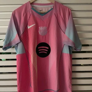 Barça x Nike rosa fotbollströja XL - Snygg FC Barcelona fotbollströja från Nike i rosa och ljusblått med vita linjer och svart Spotify-logga på bröstet. Tillverkad i Dri-FIT-material som andas. Kortärmad modell med klubbmärke och UNHCR-tryck på ryggen. Perfekt för dig som vill sticka ut på planen.