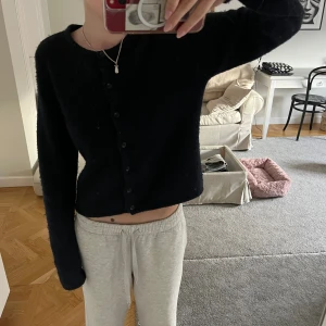 Svart stickad kofta med knappar - Mysig svart stickad kofta med rund hals och knappar framtill. Modellen är croppad och har långa ärmar, perfekt för att ge en chill och stilren vibe. Passar dig som gillar enkel och klassisk stil med en twist.