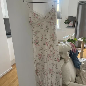 Lång blommig klänning med spetsdetaljer - Romantisk långklänning i ljusbeige med rosa blommigt mönster och tunna axelband. Klänningen har v-ringning och spetsdetaljer längs urringningen och axelbanden. Materialet är glansigt och känns svalt, perfekt för sommarens varma dagar.