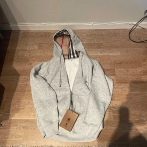 Grå Burberry hoodie med rutig huva - Snygg grå hoodie från Burberry i storlek M. Hoodien har dragkedja, snörning och den klassiska Burberry-rutiga insidan på huvan. Tillverkad i mjuk bomull, perfekt för chill dagar. 