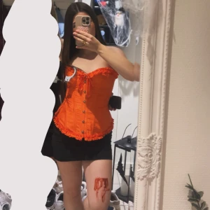 Orange korsett med volangdetaljer - Säljer en snygg orange korsett med volangkant upptill och nedtill. Toppen har knäppning framtill med metallknappar och snörning vid bysten. Materialet är glansigt och har ett diskret mönster. Perfekt för dig som vill sticka ut med en färgstark look. Den är också helt ny bara använt den 1 gång. 