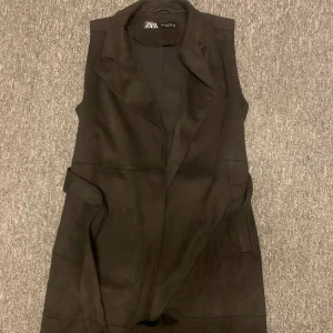 Svart lång väst från Zara i mocka - Stilren svart lång väst från Zara med breda slag och snyggt skräddad passform. Västen är ärmlös och har fickor i sidorna. Tillverkad i mjuk mocka-look som ger en lyxig känsla och passar perfekt för lager-på-lager outfits.