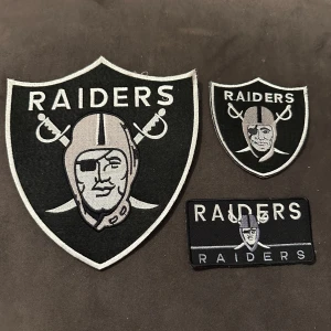Raiders tygmärke svart och silver - Säljer tre olika tygmärken med Raiders-logga i svart och silver. Märkena har sköldform och rektangulär form, med broderade detaljer och texten 'RAIDERS' samt korsade svärd. Perfekt att sy fast på keps eller mössa för en sportig look. Stort märke 19x21 cm 100kr. Det mindre 10x10cm 60kr. Det fyrkantiga 6x12 cm 60kr.