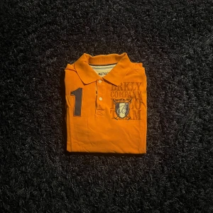 Orange pikétröja från Berkeley - Snygg orange pikétröja från Berkeley med klassisk krage, knappar och tryck på bröstet. Tröjan har en stor siffra 1 och ett coolt emblem framtill. Perfekt för dig som gillar sportig och preppy stil.