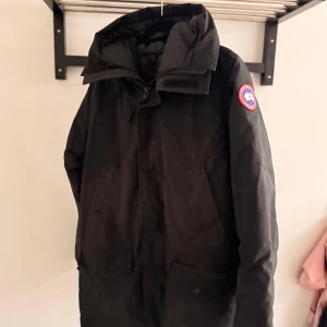 Svart dunjacka från Canada Goose - Svart dunjacka från Canada Goose med stor huva och klassisk logga på ärmen. Jackan har två stora fickor framtill och dragkedja med vindslå. Perfekt för kalla dagar och har en clean, stilren look som funkar till allt. Såklart äkta.
