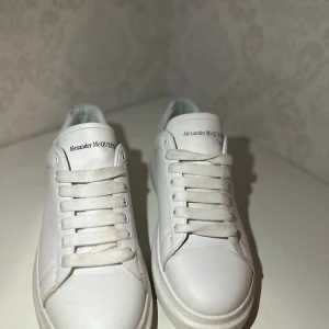 Vita sneakers från Alexander McQueen - Säljer ett par klassiska vita sneakers från Alexander McQueen med chunky sula och stilren design. Skorna har rund tå, snörning och är tillverkade i skinn. Perfekta för dig som gillar minimalistisk och trendig stil.