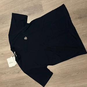 Svart t-shirt från Moncler med logga - Svart t-shirt från Moncler med klassisk rund hals och korta ärmar. På bröstet sitter en snygg Moncler-logga i svart, vitt och rött. T-shirten är gjord i mjuk bomull och har en clean, stilren look som funkar till allt.