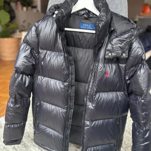 Svart pufferjacka från Polo Ralph Lauren - Svart pufferjacka från Polo Ralph Lauren med röd broderad logga på bröstet. Jackan har huva, dragkedja framtill och två sidofickor med dragkedja. Materialet är glansigt och syntetiskt, perfekt för kalla dagar. Klassisk och stilren look.