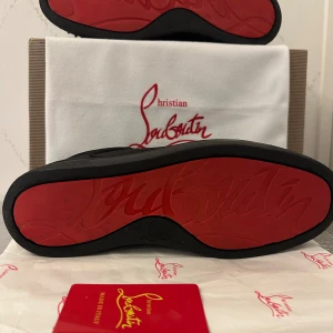 Christian Louboutin svarta sneakers med nitar - Säljer ett par svarta sneakers från Christian Louboutin med ikonisk röd sula och coola svarta nitar på tån. Skorna har snörning och är tillverkade i mocka för en lyxig känsla. Perfekta för dig som vill sticka ut med en edgy och exklusiv look.
