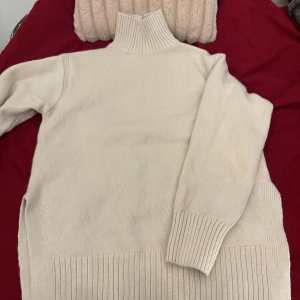 Beige stickad polotröja - Snygg beige stickad polotröja med hög krage och långa ärmar. Tröjan har breda ribbade muddar vid hals, ärmslut och nederkant som ger en schysst och modern look. Perfekt för dig som gillar stilrena och mysiga plagg.