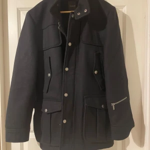 Svart rock från Blueprint med fickor - Svart rock från Blueprint med snygga detaljer som silverfärgade knappar och dragkedja på ärmen. Jackan har flera stora fickor rfärgade knappar och dragkedja på ärmen. Jackan har flera stora fickor framtill och en klassisk krage. Materialet är ull och insidan är quiltad för extra värme. Perfekt för höst och vinter.