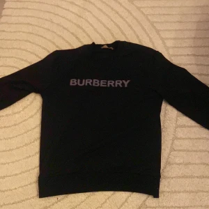 Svart sweatshirt från Burberry - Svart sweatshirt från Burberry i storlek S med klassisk logotyp tryckt över bröstet. Tröjan har rund hals, långa ärmar och ribbade muddar vid ärmslut och nederkant. Perfekt för dig som gillar stilrena och exklusiva plagg.