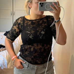 Svart spetsblus med blommönster - Säljer en svart, transparent spetsblus med blommigt mönster och korta ärmar. Toppen har en dragkedja i ryggen men är för liten och sitter tight mot kroppen. Perfekt för dig som gillar att sticka ut med coola detaljer och älskar blommig spets. 