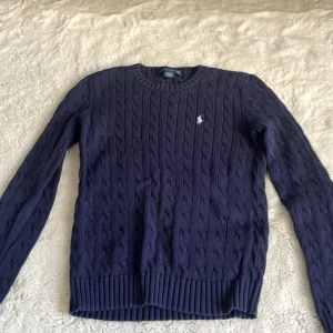 Mörkblå kabelstickad tröja Ralph Lauren - Kabelstickad mörkblå tröja från Ralph Lauren i 100% bomull. Klassisk rund halsringning, ribbade muddar och diskret broderad logga på bröstet. Perfekt för dig som gillar stilren och tidlös design.