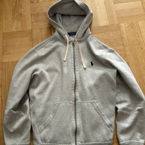 Ralph lauren hoodie - Snygg ljusgrå hoodie från Polo Ralph Lauren med dragkedja och klassisk logga broderad på bröstet. Tröjan har huva med snörning, känguruficka och är tillverkad i mjuk bomullsmix. Perfekt för chill dagar eller när du vill ha en clean look.