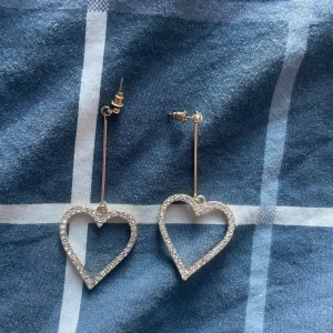 Glittriga hjärtformade örhängen - Säljer ett par långa örhängen i guld med stora hjärtan längst ner. Framsidan är täckt av glittrande stenar som ger extra bling, medan baksidan är slät och enkel. Perfekta för dig som vill sticka ut med en söt och trendig accessoar.