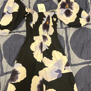 Blommig kortklänning med puffärm - Superfin kortklänning med stora blommor i svart, vit och lila. Klänningen har puffiga kortärmar, smockad överdel och offshoulder-modell. Tillverkad i ett skönt linnematerial, perfekt för varma dagar. Snygg och trendig look med voluminösa ärmar och markerad midja.