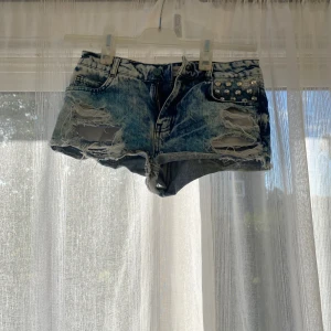 Blå jeansshorts med nitar - Säljer ett par blå jeansshorts med coolt slitna detaljer och råa kanter. Shortsen har nitar på ena fickan och en klassisk knapp och dragkedja framtill. Perfekt för dig som gillar edgy streetstyle och vill sticka ut. Passar XS/S