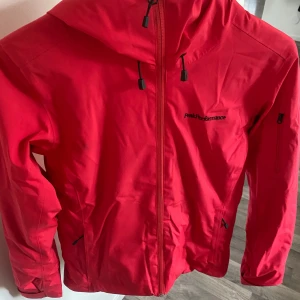 Röd vindjacka Peak Performance XS - Snygg röd vindjacka från Peak Performance med huva och svarta detaljer. Jackan har dragkedja framtill, praktiska fickor och justerbara snören vid huvan. Insidan är svart och jackan är tillverkad i slitstark polyester, perfekt för blåsiga dagar.