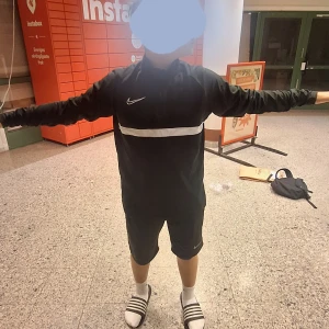 Nike svart Dri-Fit half zip tröja M - Svart långärmad Nike Dri-Fit tröja med vit rand över bröstet och broderad Swoosh-logga. Tröjan har half zip och är gjord i ett lätt, syntetiskt material som andas. Perfekt för träning eller chill. Sitter normalt och har sportig vibe.