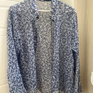 Blåvit blommig skjorta i tunn polyester - Säljer en blåvit skjorta med blommigt mönster i tunn och luftig polyester. Skjortan har långa ärmar, klassisk krage och knäppning Blåvit blommig skjorta i tunn polyester. Perfekt för dig som gillar en lätt och somrig vibe med snyggt blommigt print.