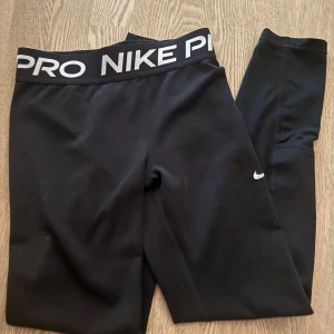 Svarta Nike Pro leggings - Svarta Nike Pro leggings med bred resår i midjan där det står NIKE PRO i vitt. Sitter tight och är perfekta för träning eller chill. Materialet är stretchigt och andas, så du kan röra dig fritt och bekvämt.