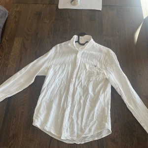 Vit långärmad skjorta i bomull - Säljer en stilren vit linne skjorta  med klassisk krage och bröstficka. Skjortan har långa ärmar och knäppning framtill. Använd några få gånger. Skrynklig orkade inte stryka den 