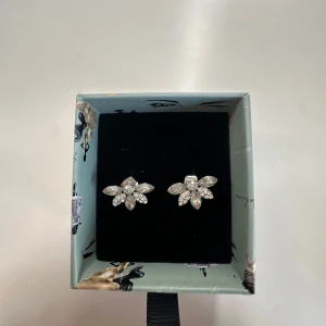 Blommiga örhängen i silverfärg - Snygga örhängen i silverfärg med blommig design, dekorerade med glittrande stenar som ger en lyxig känsla. Perfekta för att lyfta din stil med lite extra bling. Kommer i en fin ask.