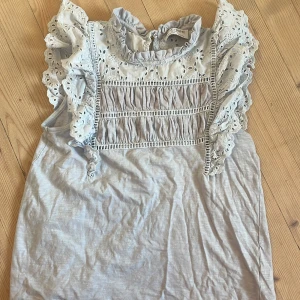 Ljusblå blus med volang från Holly & Whyte - Supersöt ljusblå blus från Holly & Whyte med broderade detaljer och volang vid axlarna. Blusen har ärmlös design, rynkade partier framtill och romantisk spets längs halsringningen. Perfekt för dig som gillar en feminin och somrig stil.