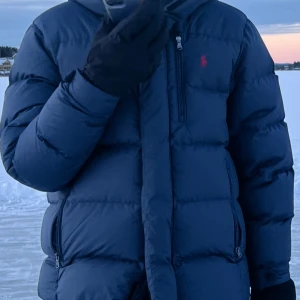Mörkblå dunjacka från Ralph Lauren - Snygg mörkblå dunjacka från Ralph Lauren med klassisk pufferstil och dragkedjor på fickorna. Jackan har en diskret röd logga på bröstet och är perfekt för kalla dagar. Fylld med dun för extra värme och har en hög krage som skyddar mot vinden.