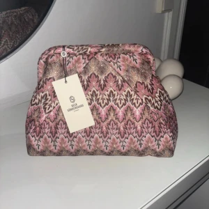 Mönstrad kuvertväska från Beck Söndergaard - Kuvertväska med unikt mönster i rosa, brunt och beige från Beck Söndergaard. Väskan har en klassisk form, mjukt tygmaterial och innerfack med dragkedja. Perfekt för att lyfta din outfit med lite extra färg och stil.