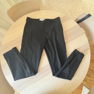 Svarta kostymbyxor från Les deux - Snygga svarta kostymbyxor från les deux med klassisk rak passform och diskreta fickor i sidorna. Perfekta för en clean och stilren look. Byxorna har hällor för bälte och stängs med dragkedja och knapp.