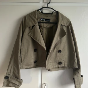 Croppad trenchcoat från Zara  - Säljer en croppad trenchcoat från Zara i beige bomullsmaterial. Jackan har dubbelknäppning framtill, breda slag och snygga detaljer med knappar vid ärmsluten. Perfekt för dig som vill ha en trendig och stilren look.