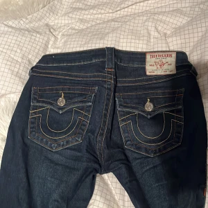 True Religion Joey Low Rise Flare Jeans - Säljer ett par mörkblå True Religion Joey jeans med låg midja och utsvängda ben. Snygga kontrastsömmar, ikoniska bakfickor med lock och silverknappar. Klassisk denim med coola detaljer och riktigt skön passform.