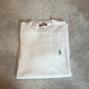 Vit kabelstickad pikétröja Polo Ralph Lauren - Vit kabelstickad pikétröja från Polo Ralph Lauren med korta ärmar och klassisk rund hals. Tröjan har det ikoniska broderade logotypen på bröstet och är gjord i mjuk bomull. Perfekt för en clean och stilren look.