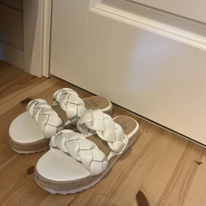 Vita flätade sandaler med platåsula - Snygga vita sandaler från Vox med två breda, flätade remmar i syntetmaterial. Platåsulan har en snygg detalj i rep och ger extra höjd. Perfekta för sommarens alla dagar och lätta att matcha med olika outfits.