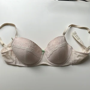 Ljusrosa spets-bh från Twilfit 70D - Söt ljusrosa bh från Twilfit i storlek 70D. Bh:n har vadderade kupor med spetsdetaljer och en liten rosett framtill. Axelbanden är justerbara och knäppningen bak har flera hakar för bästa passform.